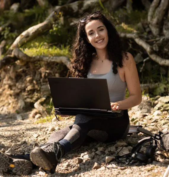Brunette woman in a grey tank top, black pants, and hiking books sitting down with a laptop in her lap
