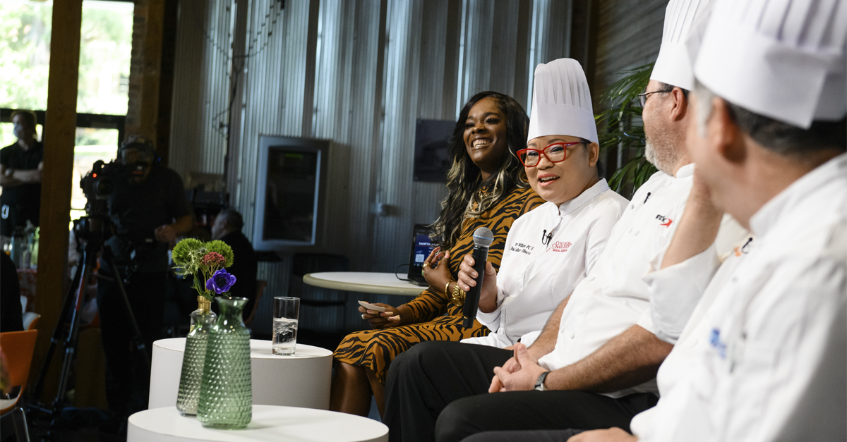 2 male chefs look at a female chef speaking as a part of a panel. The panel facilitator looks out into the audience.