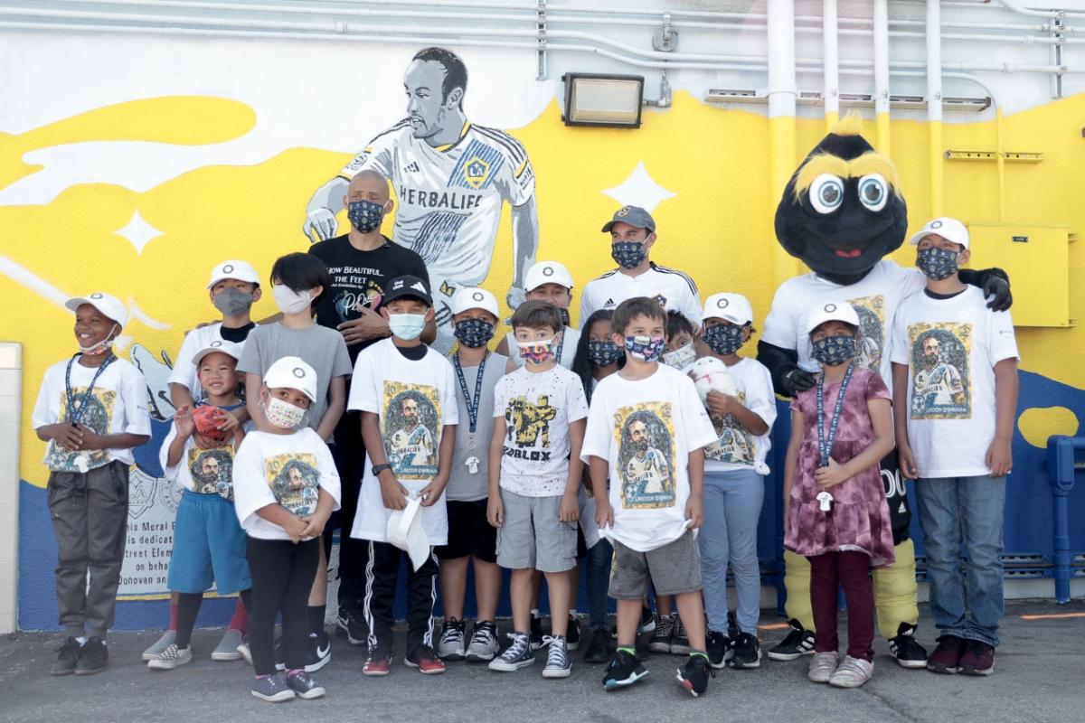 Students in Landon Donovan t-shirts and masks crowd around LA Galaxy player Landon Donovan in front of a yellow, white and blue mural of Landon Donovan in an LA Galaxy jersey. 