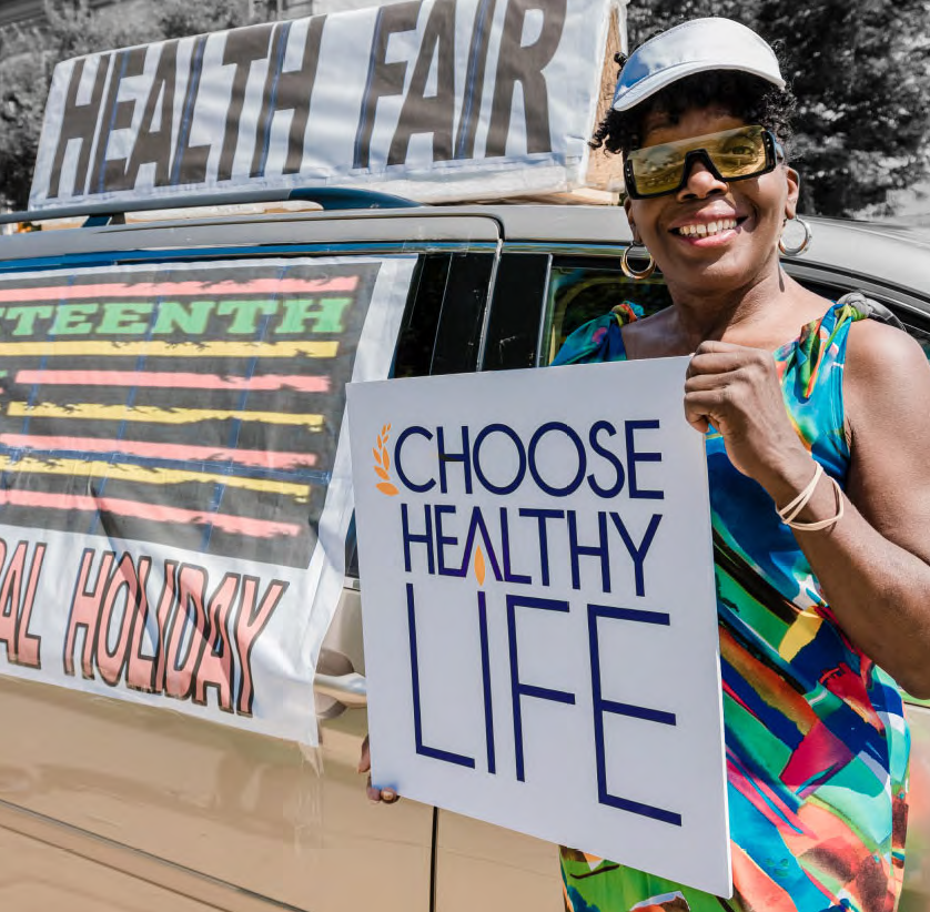 a person holds a sign "choose healthy life" infront of a vehicle with "health fair" on the roof and a sign over the window "...teenth...Holiday"