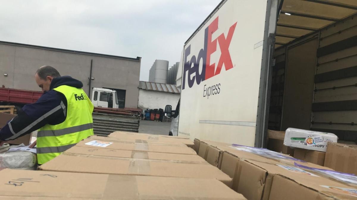 view from the top of a row of boxes towards a person checking them. A fedex truck to the right