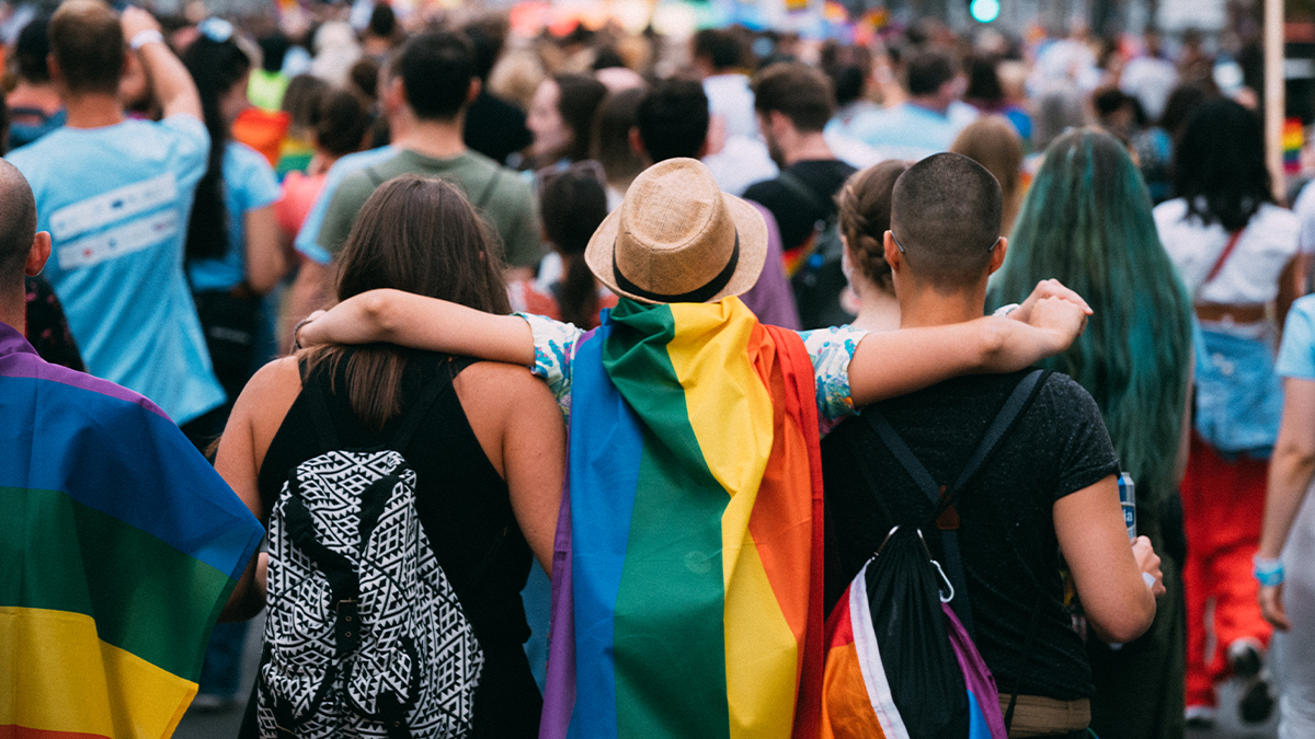 Person with rainbow flag cape embraces two other people